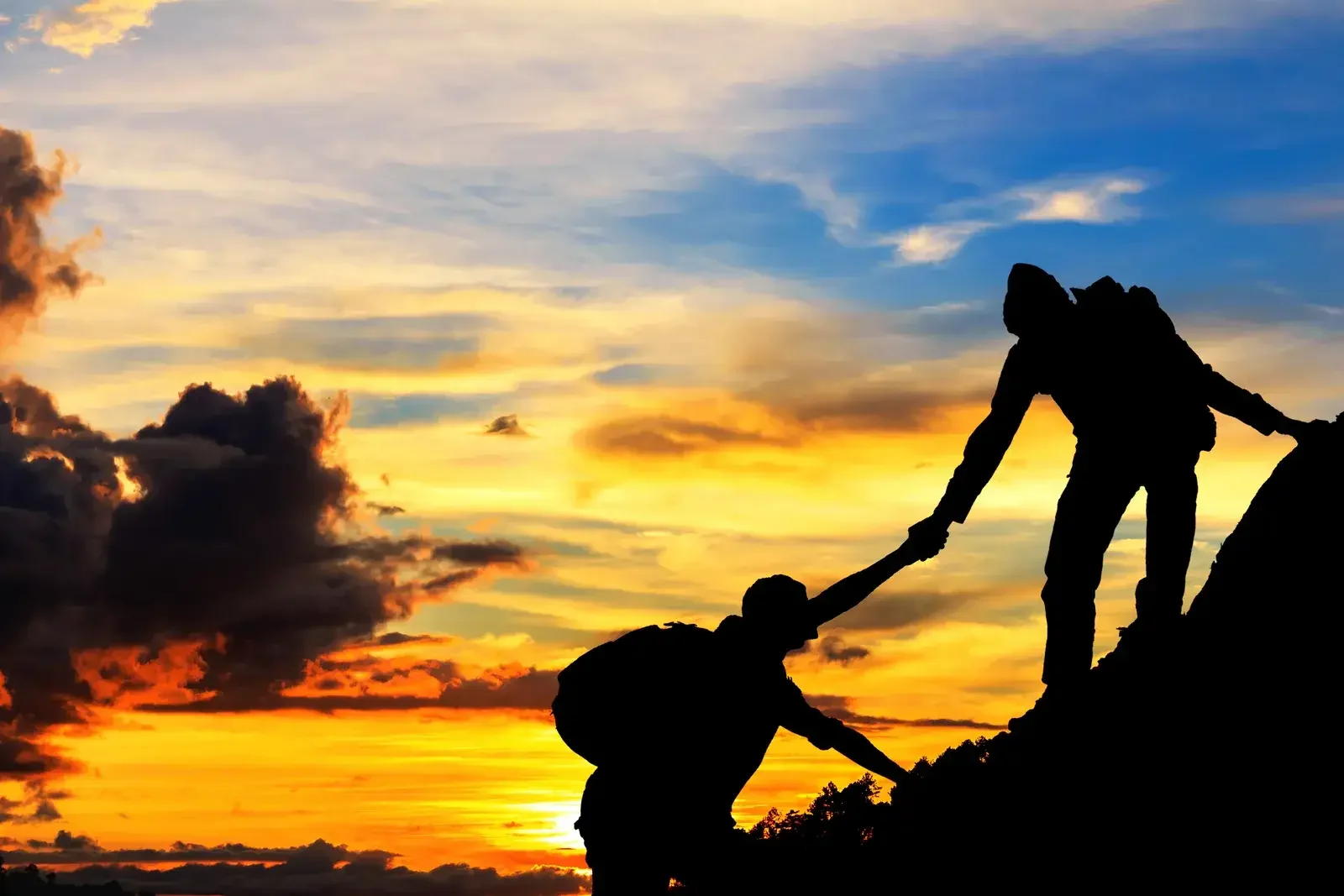 Silhouettes de deux randonneurs sur une crête rocheuse, l’un tendant la main à l’autre pour l’aider à monter, devant un ciel de coucher de soleil orangé