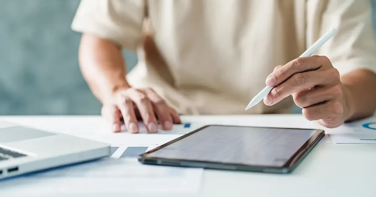 Main tenant un stylet au-dessus d'une tablette posée près d'un ordinateur portable, sur un bureau clair