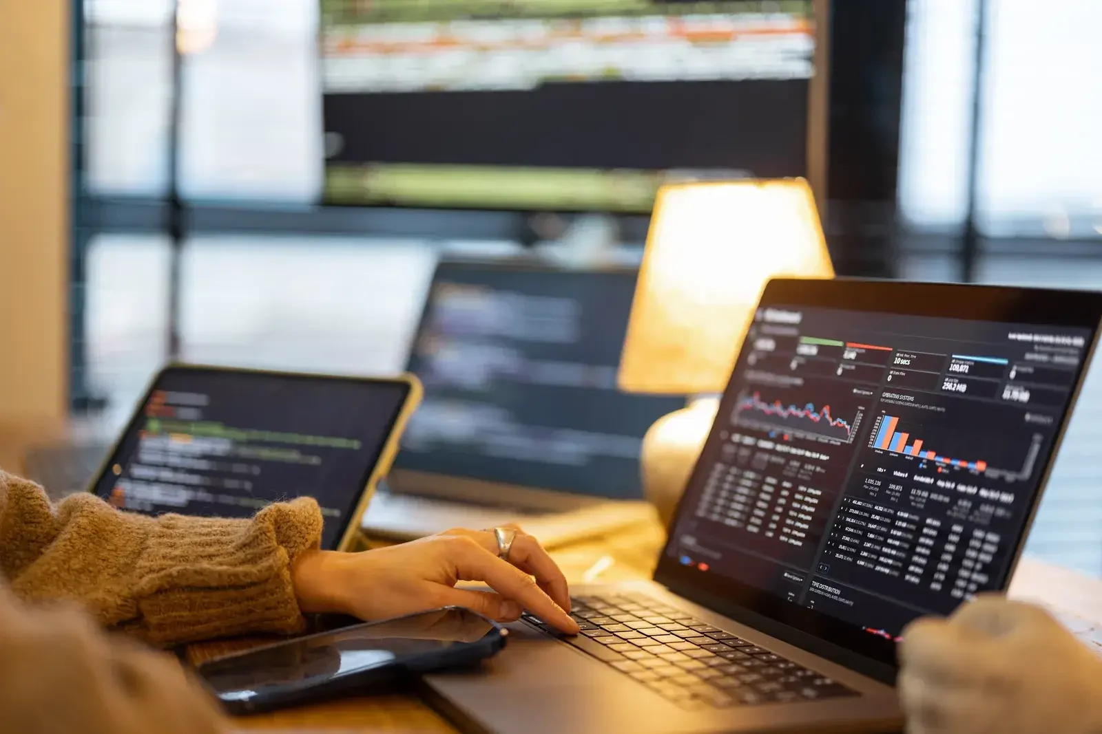 Mains d’une personne en pull beige sur le clavier d’un ordinateur portable, plusieurs écrans affichant des graphiques et des tableaux de bord, lampe à lumière chaude en arrière-plan