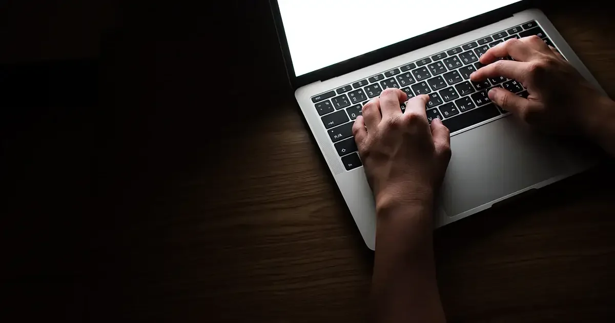 Mains posées sur le clavier d'un ordinateur portable dans une pièce sombre, lumière chaude rasante sur le bois