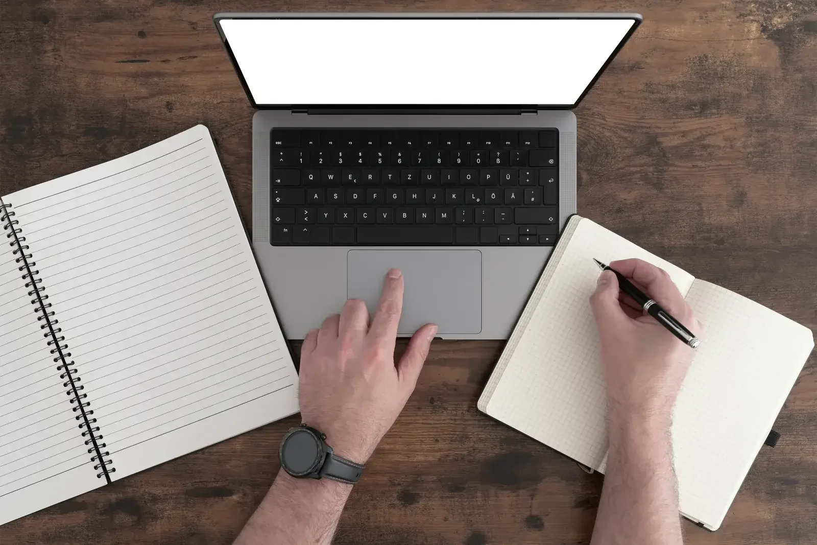 Vue de dessus d’une table en bois foncé : une main sur le clavier d’un ordinateur portable, l’autre écrivant dans un carnet ouvert, un bloc à spirale posé à côté